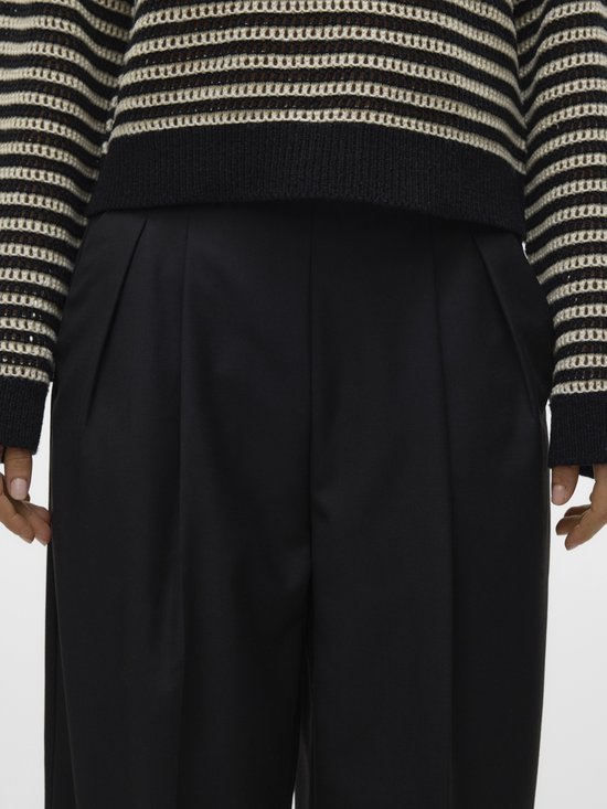 Close-up van een zwarte wijde broek met plooien aan de voorkant, gedragen met een gestreepte trui.
