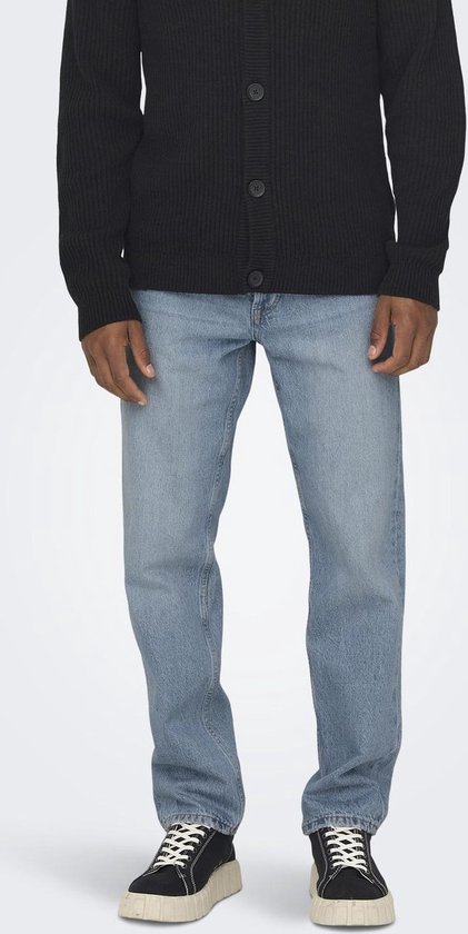 Man staat, draagt een zwarte cardigan en lichtblauwe jeans, met witte sneakers.