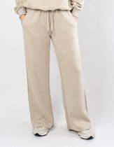 Een beige joggingbroek met wijde pijpen en trekkoord, gedragen door een model. Logo op linker dij.