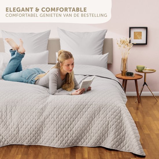 Vrouw leest op een lichtgrijs, gewatteerd bed. De sprei heeft een ruitpatroon.