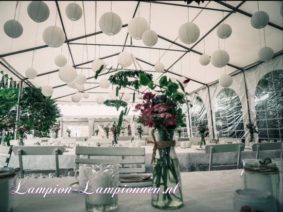 Feesttent met witte lampionnen, gedekte tafels en bloemstukken.