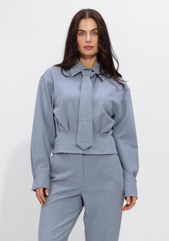 Model in een lichtblauwe blouse met strik en bijpassende pantalon. De blouse heeft een ceintuurdetail.