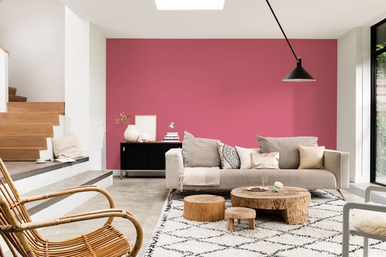 Een woonkamer met een roze muur, een beige bank met kussens, een rotan stoel en houten tafels.
