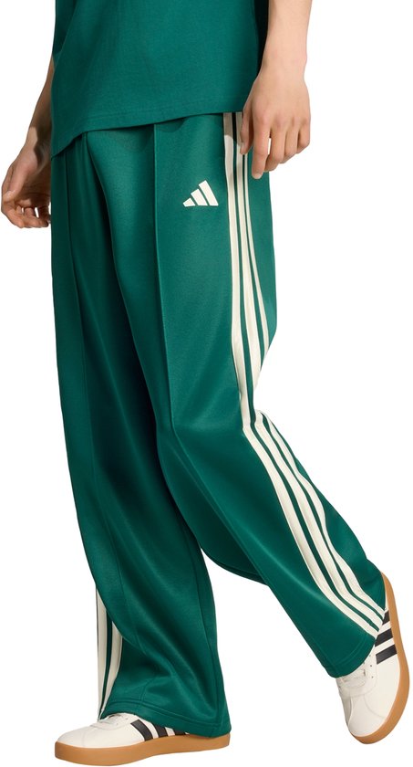 Een model draagt een donkergroene wijde broek met beige Adidas-strepen en witte sneakers.