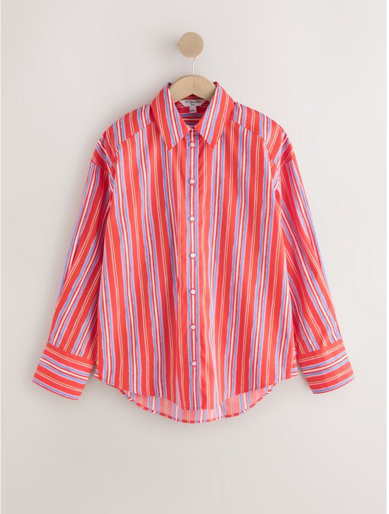 Oversized rood overhemd met verticale blauwe, witte en roze strepen, hangend aan een kledinghanger.