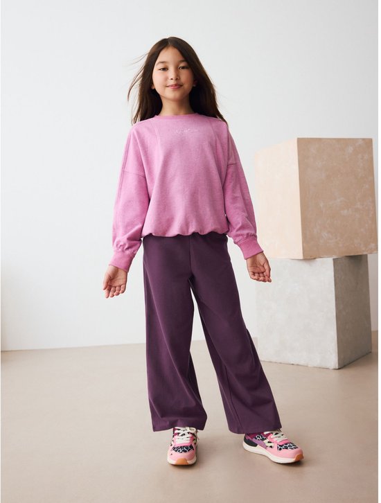 Staand kind in een roze trui en een paarse wijde tricot broek.