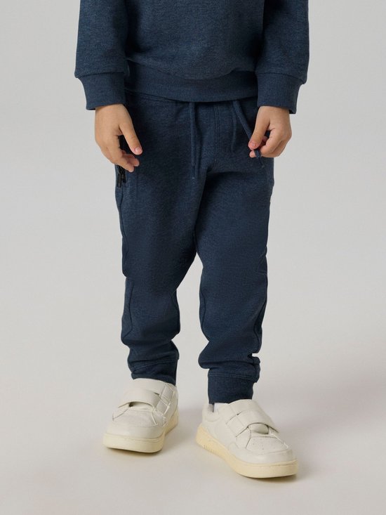 Onderlichaam van een jongen in een donkerblauw joggingbroek en witte sneakers.