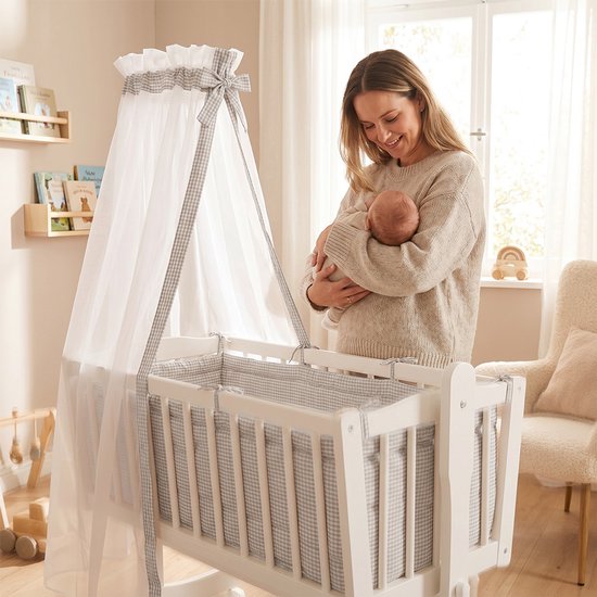 Wit kinderbed met hemel, ruitjespatroon en vrouw met baby.