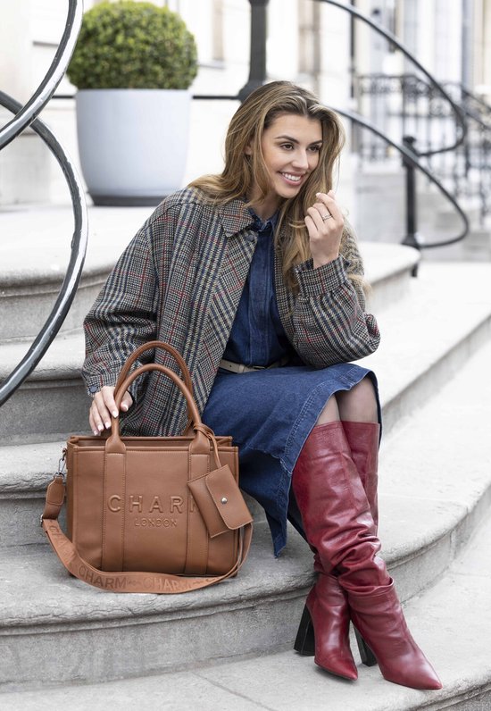 Model met bruine tote bag met 'CHARM LONDON' in reliëf en bijpassend etui, gedragen met een geblokte jas en rode laarzen.