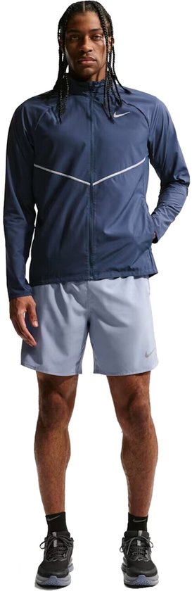 Man in blauw Nike hardloopjack met zilveren streep, Nike logo, en blauwe shorts. Staat met handen in de zakken.