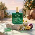 Groene Nuxe Huile Prodigieuse Néroli fles met gouden dop, op stenen met pruimen en witte bloesems.