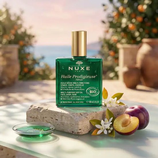 Groene Nuxe Huile Prodigieuse Néroli fles met gouden dop, op stenen met pruimen en witte bloesems.