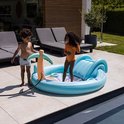 Kinderen spelen in een opblaasbaar zwembad met palmtak sproeier en glijbaan. Een hose is aangesloten op de sproeier.