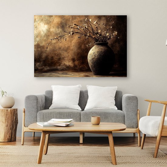 Canvasafdruk met een donkere, aardse vaas gevuld met gedroogde bloemen. Achtergrond is een bruin en goudkleurig abstract schilderij. Hangt boven een grijze bank.