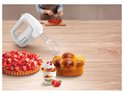 Witte Tefal handmixer op een houten tafel naast gebak en fruitdessert, met een persoon op de achtergrond.