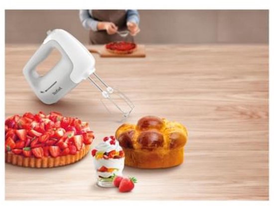 Witte Tefal handmixer op een houten tafel naast gebak en fruitdessert, met een persoon op de achtergrond.