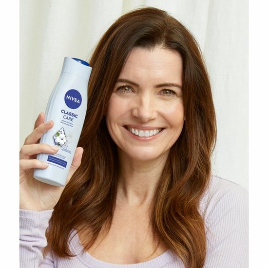 Vrouw met bruin haar lacht en houdt een fles NIVEA Classic Care shampoo omhoog, met afbeelding van een katoenbloem.