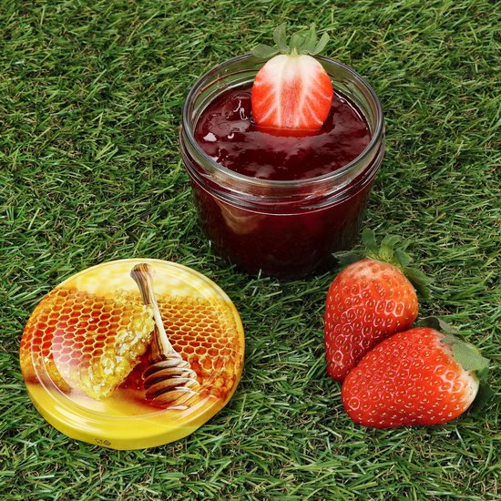 Glazen pot met rode jam, versierd met een aardbei, met honingraatdeksel ernaast, op gras.