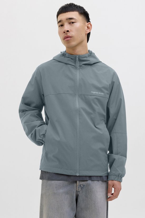 Model draagt een zilvergrijze jas met capuchon en spijkerbroek. De jas heeft een ritssluiting en de handen in de zakken.