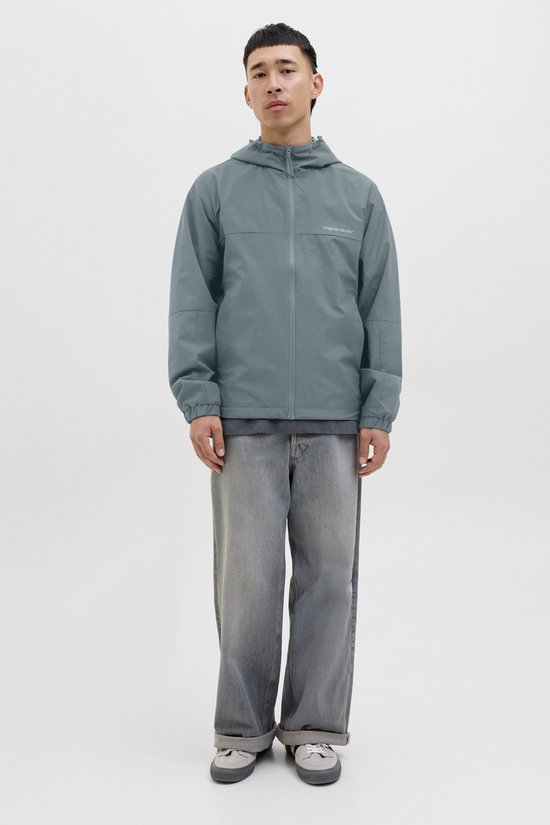 Model draagt een zilvergrijze jas met capuchon en spijkerbroek. De jas heeft een ritssluiting en manchetten met elastiek.