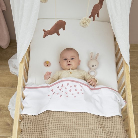 Baby in een bedje met een wit dekentje met roze hartjes.