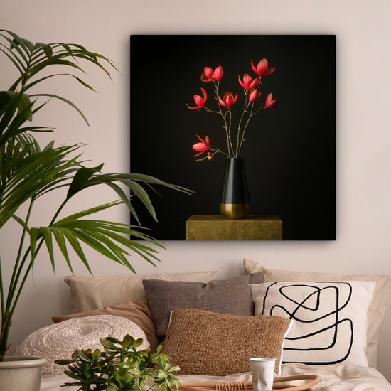Rode bloemen in een zwarte vaas op een gouden sokkel, in een slaapkamer met planten en kussens.