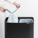 Poubelle Brabantia noire avec deux compartiments ouverts contenant des sacs poubelles bleu clair. Deux mains ferment les sacs par le haut.