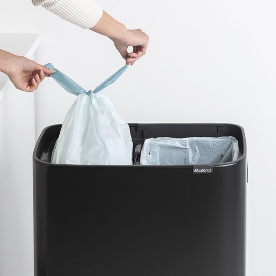Poubelle Brabantia noire avec deux compartiments ouverts contenant des sacs poubelles bleu clair. Deux mains ferment les sacs par le haut.