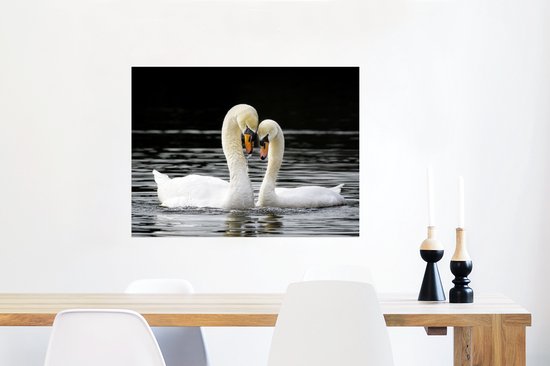 Deux cygnes blancs au bec orange nagent dans l'eau sombre, leurs cous forment un cœur, accrochés au mur au-dessus d'une table.