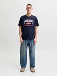 Man in marineblauw T-shirt met wit-rode 'JACK & JONES' print en wijde spijkerbroek.