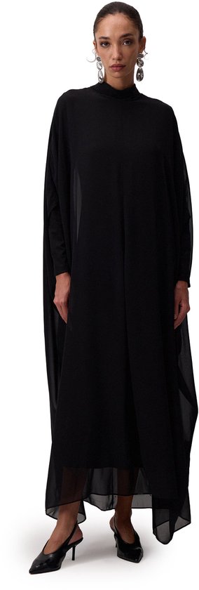 Model draagt een zwarte chiffon jurk met hoge hals en wijde vleermuismouwen, gecombineerd met zwarte hakken.