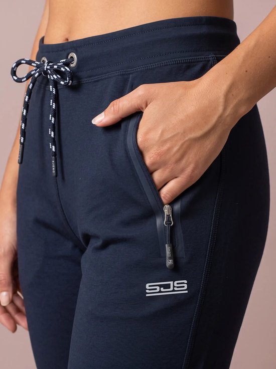 Close-up van marineblauwe sportbroek met een ritszak en het Sjeng logo, hand in de zak gestoken.