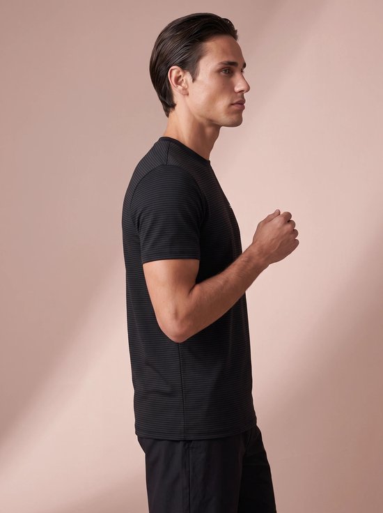 Zijaanzicht van een man in een zwart, fijn gestreept sportshirt met korte mouwen, staand tegen een neutrale achtergrond.