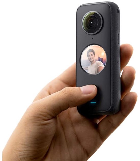 Hand met zwarte Insta360 One X2 camera, rond scherm toont man met lach, zonsondergang zichtbaar.