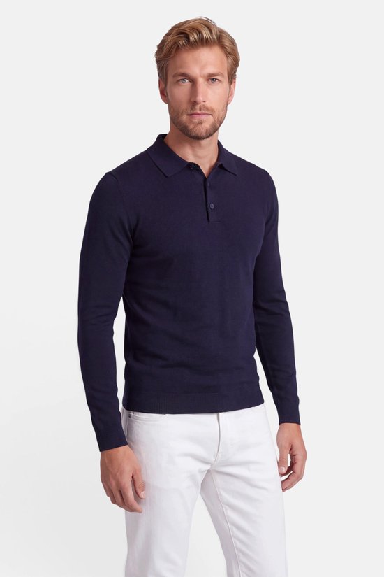 Model in een marineblauwe polo met lange mouwen en een witte broek.