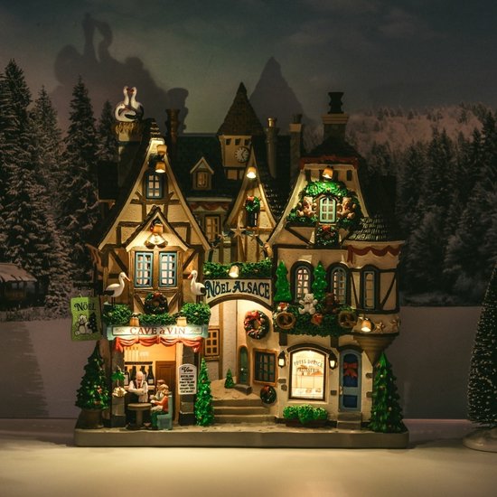 Verlichte Noël Alsace kerstminiatuur in het donker, met warme glans vanuit de wijnbar en patisserie.