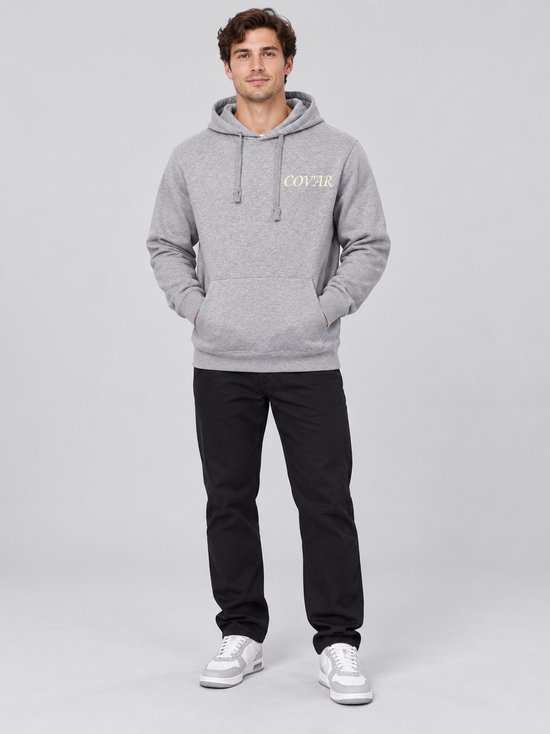 Man met grijze hoodie met 'COVAR' print op linkerborst. Handen in zakken.