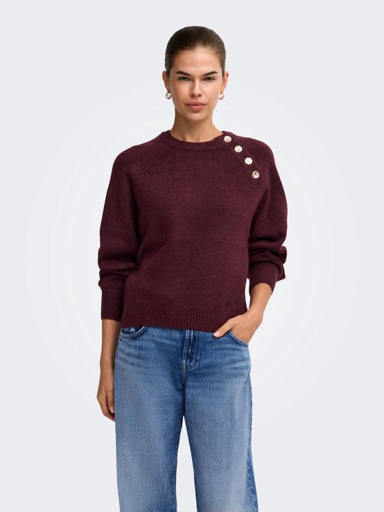 Model met een wijnrode gebreide trui met vier gouden knopen op de schouder en jeans.