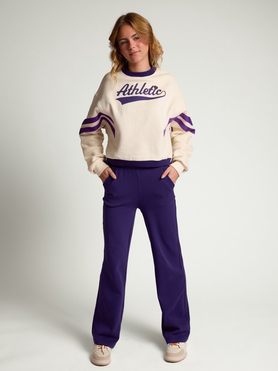Model draagt een ecru sweater met paarse accenten en 'Athletic' tekst, gecombineerd met een paarse broek.