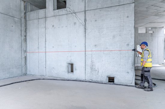 Bouwvakker in werkkleding gebruikt een rode laser-afstandsmeter in een onafgewerkte betonnen ruimte.