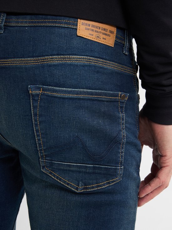 Detail van achterkant donkerblauwe spijkerbroek met stonewash en het label 'DENIM DRIVEN SINCE 1989'.