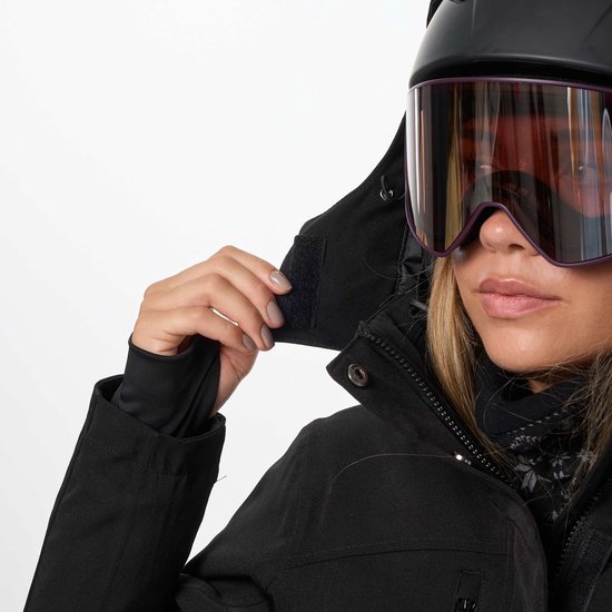 Vrouw sluit de capuchon van haar zwarte ski-jas, met paarse skibril en helm.