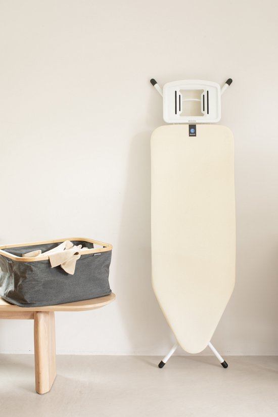 Crèmekleurige strijkplank rechtop met ophangbeugel, naast een grijze wasmand op een houten tafel.