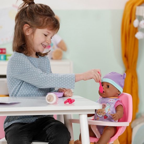 Meisje voert babypop in een kinderstoel aan tafel. De pop draagt een paarse muts en rompertje.