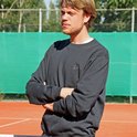 Man met donkerblauwe trui en armen gekruist, staand bij een tennisbaan.