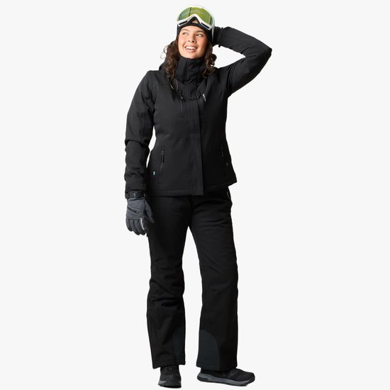 Vrouw draagt zwarte ski-jas en broek, met ski-bril en handschoenen. Ziet er blij uit.