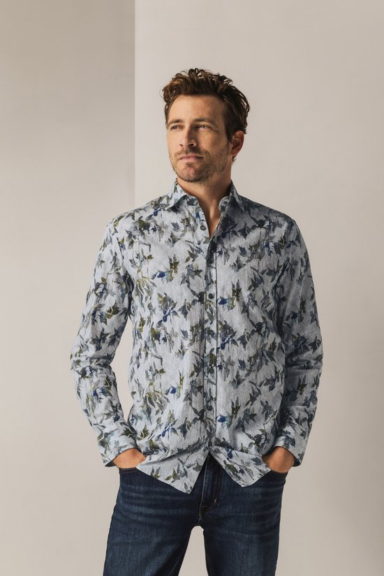 Man met handen in de zakken kijkt weg, draagt een lichtgrijs overhemd met botanische print.