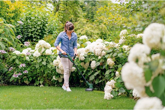 Vrouw trimt gras met Bosch EasyGrassCut 18V-260 grastrimmer in een tuin met witte hortensia's.