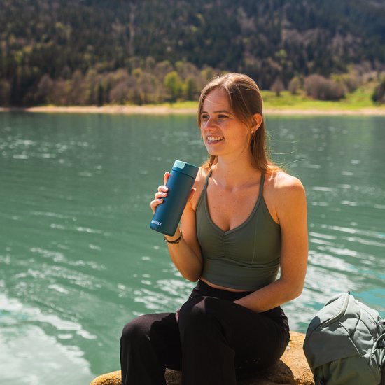 Vrolijke vrouw met een blauwe Kambukka thermosbeker bij een meer.