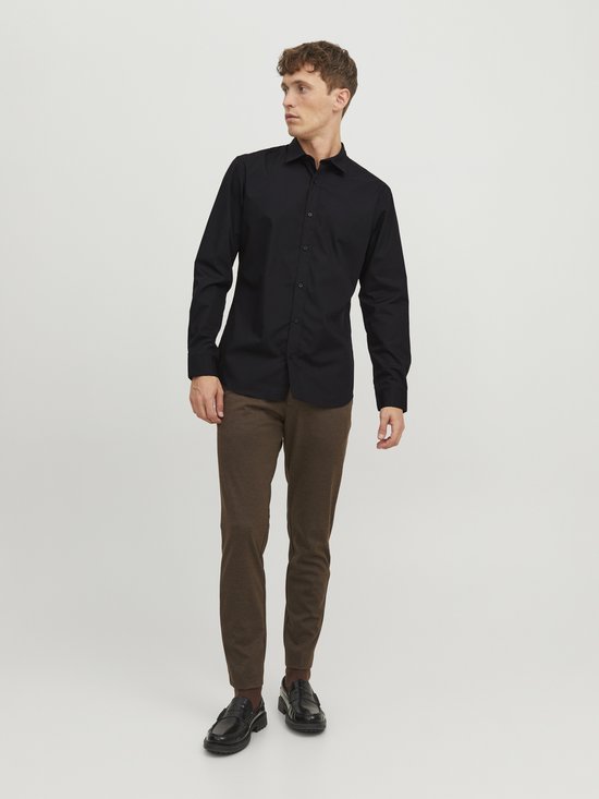 Man in zwart, lang-mouwig button-down overhemd en bruine broek, staand.